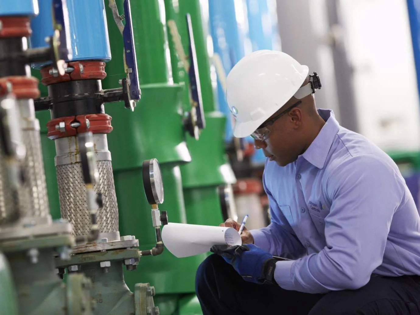 Mechanic wearing a hard hat recording equipment readings beside industrial piping and gauges